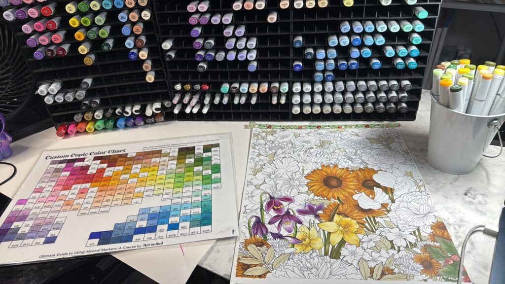 a color chart next to a coloring project of flowers on a desk.